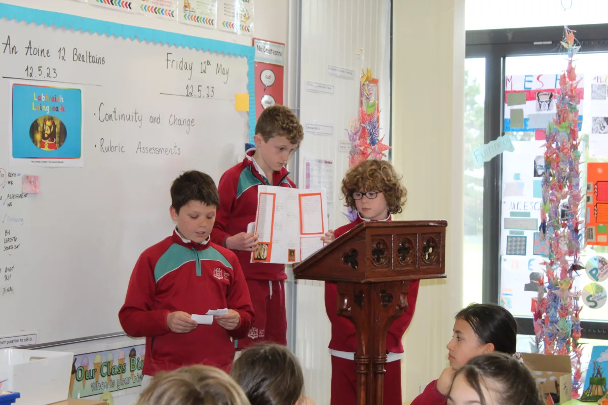 A picture of three pupils standing at the top of their classroom reading their group project to the visiting Grandparents and Special Persons in their classroom